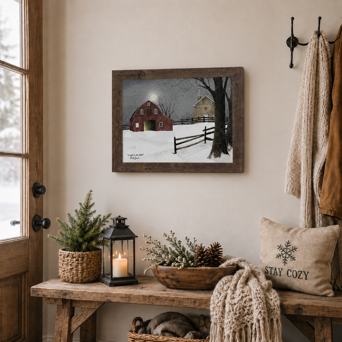 Natural brown framed Billy Jacobs "A Light in the Stable" landscape print displayed above a rustic wooden entryway bench with a lit lantern, pine tree in a basket, pinecone bowl, and a Stay Cozy pillow with coats hanging on wall hooks beside a snowy window