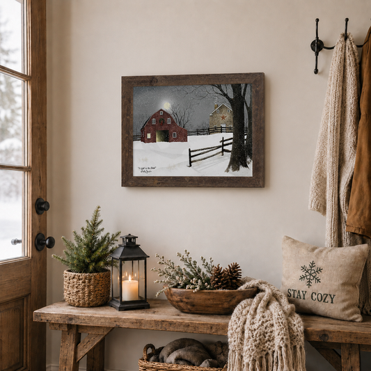 Natural brown framed Billy Jacobs "A Light in the Stable" landscape print displayed above a rustic wooden entryway bench with a lit lantern, pine tree in a basket, pinecone bowl, and a Stay Cozy pillow with coats hanging on wall hooks beside a snowy window