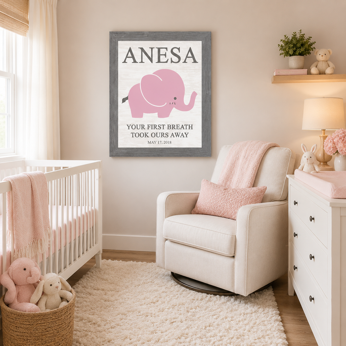 Gray framed personalized baby name nursery wall art PER149 displayed large in soft pink girl nursery above glider chair beside white crib — pink elephant with bold name ANESA at top and Your First Breath Took Ours Away with birth date May 17 2018 on soft white background — stuffed bunnies changing table and white dresser in room
